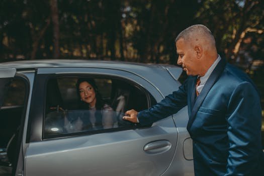 A businessman in a suit opens a car door for a woman sitting inside under sunlight outdoors.