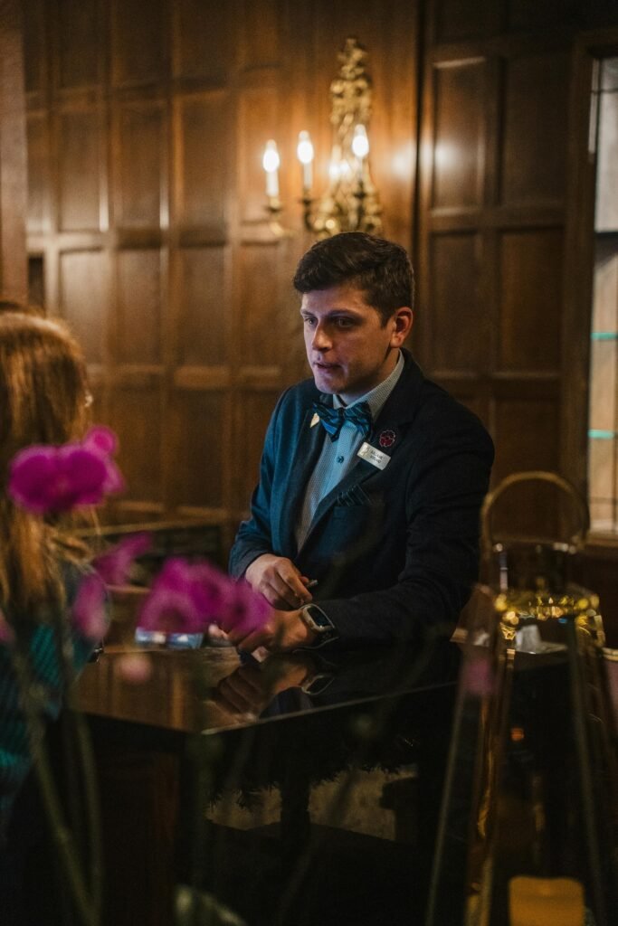 Polite hotel receptionist in formal attire at a warmly lit wooden reception desk.